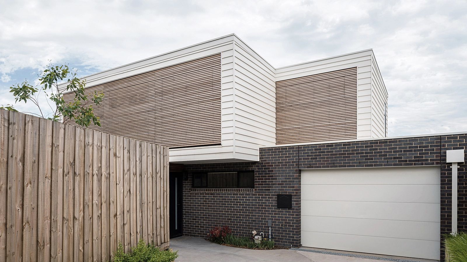 Image of an architect designed new build house.The brick floor is blue-grey brick and the first floor with weatherboard. A timber screen spans the full width of the first floor facade protecting the windows from sun exposure.